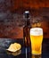 Glass with freshly poured unfiltered light beer, beer bottle near scattered chips on a black mirror surface