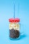 Glass jar with handle, full, closed. In the jar, coffee beans down, popcorn up. Red lid, white dots. Straw blue-white. Blue