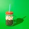 Glass jar with handle, red lid, white dots, white straw, blue stripes. In the jar, coffee beans and popcorn. Harsh shadow. Green
