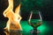 Glass o whisky with ice and wineglass of cognac and fire flames on green bokeh background