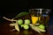 A glass with olive oil stands on a wooden table against a dark background. Next to it is a branch with olives and leaves on it