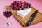 Glass of red wine and a triangular piece of Brie cheese, red sweet grapes, chef knife on a brown wooden cutting board. Soft cheese