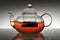 Glass teapot with hot tea on gray gradient background with reflection