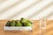 A glass of water and some lemons on the table.  Some lemons are placed on a white wooden plate