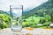 Glass of water on the stone. Blurred snow mountains tops and green forests at the background