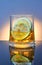 A glass of whiskey with ice and lemon on a blue background with orange backlight and reflection