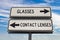 Glasses versus contact lenses road sign with two arrows on blue sky background. White two street sign with arrows on metal pole.