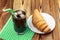 Glassful of black coffee with ice and tubule on green napkin. croissant, wooden table in cafe.