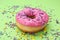 Glazed donut decorated with sprinkles on green background, closeup. Tasty confectionery