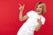 Gleeful young blonde woman in casual white outfit pointing fingers away over red background.