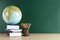 Globe, school supplies and books on wooden table near green chalkboard, space for text. Geography lesson