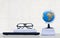 Globe on stacked books with eyeglasses and black laptop on glass tabletop with white brick wall background, front view with copy