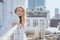 Glorious woman in white light gown with embroidery standing on rooftop leaning on fence and touching face with hand.