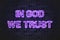 In God We Trust, the official motto of the United States glowing violet neon letters on a black brick wall