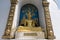 A golden statue of Buddha sitting in the lotus position, located in a niche surrounded by traditional Buddhist designs