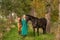 In the golden sun a portrait of a blond smiling girl with her horse and foal in the forest. Wearing a green dress. Selective focus