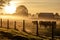 Golden sunrise over a misty farm with cows behind a rustic fence, a serene pastoral scene