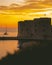 Golden sunset in the town of Dubrovnik, castle standing on the shore of a small harbour, sailboat leaving the harbour and a small