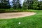 Golf ball in the grass rough in front of a sand trap, green and flag behind hazard