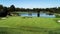 Golf course fairway, water hazard pond, sand bunkers, green with flag, lush green grass surrounded by trees against blue sky