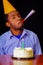 Good looking man wearing blue shirt and hat sitting by table with cake in front, single candle burning, blowing party