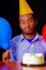 Good looking man wearing blue shirt and hat sitting by table with cake in front, single candle burning, holding balloons