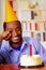 Good looking man wearing blue shirt and hat sitting by table with cake in front, single candle burning, skeptically