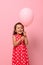 Gorgeous birthday baby girl rejoices looking at pink balloon in her hands, happily smiles posing on pink background with copy