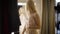 Gorgeous blond Caucasian woman in beige dress looking in big mirror in fashion boutique. Beautiful young shopper trying