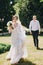 Gorgeous bride in amazing gown and stylish groom running and laughing in sunny park. Beautiful happy wedding couple enjoying time