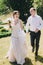 Gorgeous bride in amazing gown and stylish groom running and laughing in sunny park. Beautiful happy wedding couple enjoying time