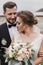 Gorgeous bride with modern bouquet and stylish groom gently hugging and smiling outdoors. Sensual wedding couple embracing. Roman