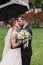 Gorgeous bride and stylish groom passionately kissing under umbrella in rainy outdoors. Sensual wedding couple embracing.