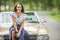 Gorgeous brunette smiles intothe camera sitting on front hood of a vintage convertible car on the roadside in teh nature