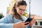 A gorgeous female in blue sweater, eyeglasses sitting on the bench reading news on smart phone outdoors on a sunny day. Happy