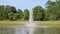 Gorgeous footage of a water fountain in the center of a silky green lake surrounded by lush green trees, grass and plants