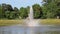 Gorgeous footage of a water fountain in the center of a silky green lake surrounded by lush green trees, grass and plants
