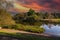 A gorgeous shot of a lake in the park surrounded by lush green and autumn colored trees and plants with powerful clouds at sunset