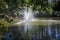 A gorgeous shot of a majestic water fountain in the middle of still lake water with lush green and autumn colored trees and plants