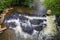 A gorgeous shot of the river water rushing over the rocks in the middle of the river with lush green trees on the banks