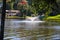 A gorgeous shot of the still blue waters of the lake with water fountain in the lake with lake front homes with lush green trees