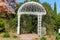 A gorgeous shot of a white rod iron gazebo surrounded by lush green grass, trees and plants near a pink and yellow tree