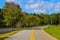 A gorgeous shot of a winding road with a yellow line through the park with lush green and autumn colored trees along the lake