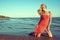 Gorgeous slim blond model in coral red strapless dress standing on knees in the sea water and looking aside