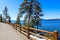 A gorgeous smooth concrete paved hiking trail with a burnt orange wooden fence along the trail with lush green pine trees and snow