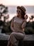 Gorgeous wealthy redhead woman at summer sunset sitting on stone parapet in sunglasses, straw hat and dress