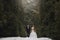 Gorgeous wedding couple posing in winter snowy park. stylish bride in coat and  groom embracing under green trees in winter forest