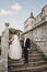 Gorgeous wedding couple walking on stone stairs near old castle in park. Stylish beautiful bride in amazing gown and  groom posing