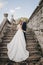 Gorgeous wedding couple walking on stone stairs near old castle in park. Stylish beautiful bride in amazing gown and  groom posing