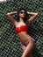 Gorgeous woman with slim bronzed body in red stylish bikini and sunglasses is posing near grid and wall of green leaves